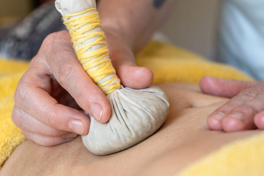 Nahaufnahme einer Hand die einen Kräuterstempel hält und sanft den Bauch einer Klientin massiert.